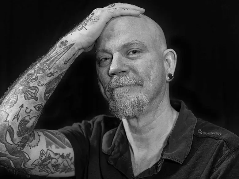 Photo of a middle-aged bald white man with a goatee. His right hand is resting on his head, exposing a densely tattooed arm.
