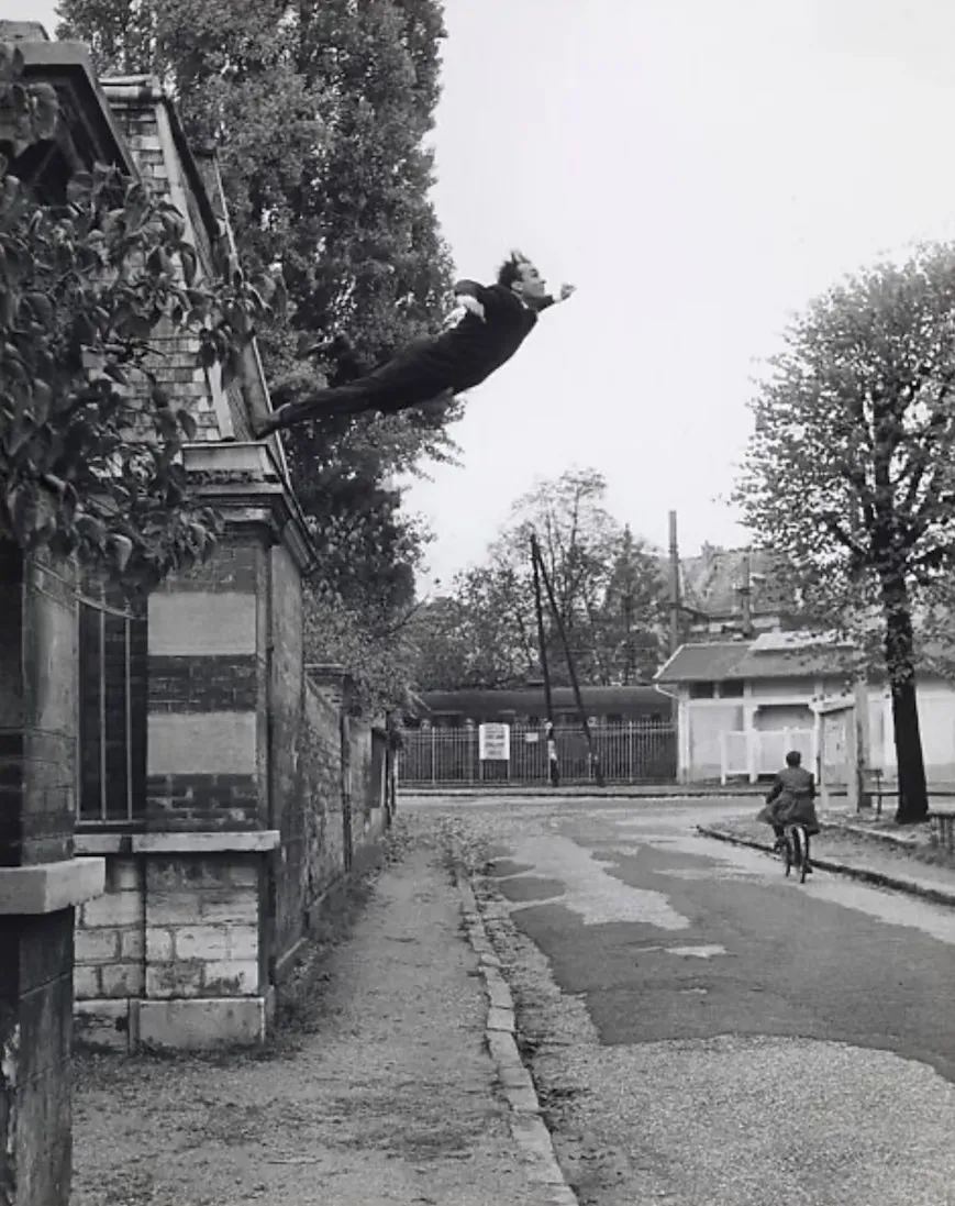 vintage black and white photo of a man in a suit doing a swan dive off a roof over the pavement of a street 