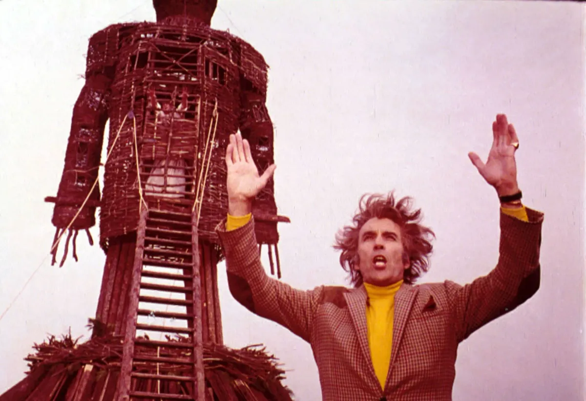 Vintage photo of a guy in a yellow turtleneck and wild hair, with arms outstretched over his head. Behind him is a giant wickerman. The mood is frenzied magic.