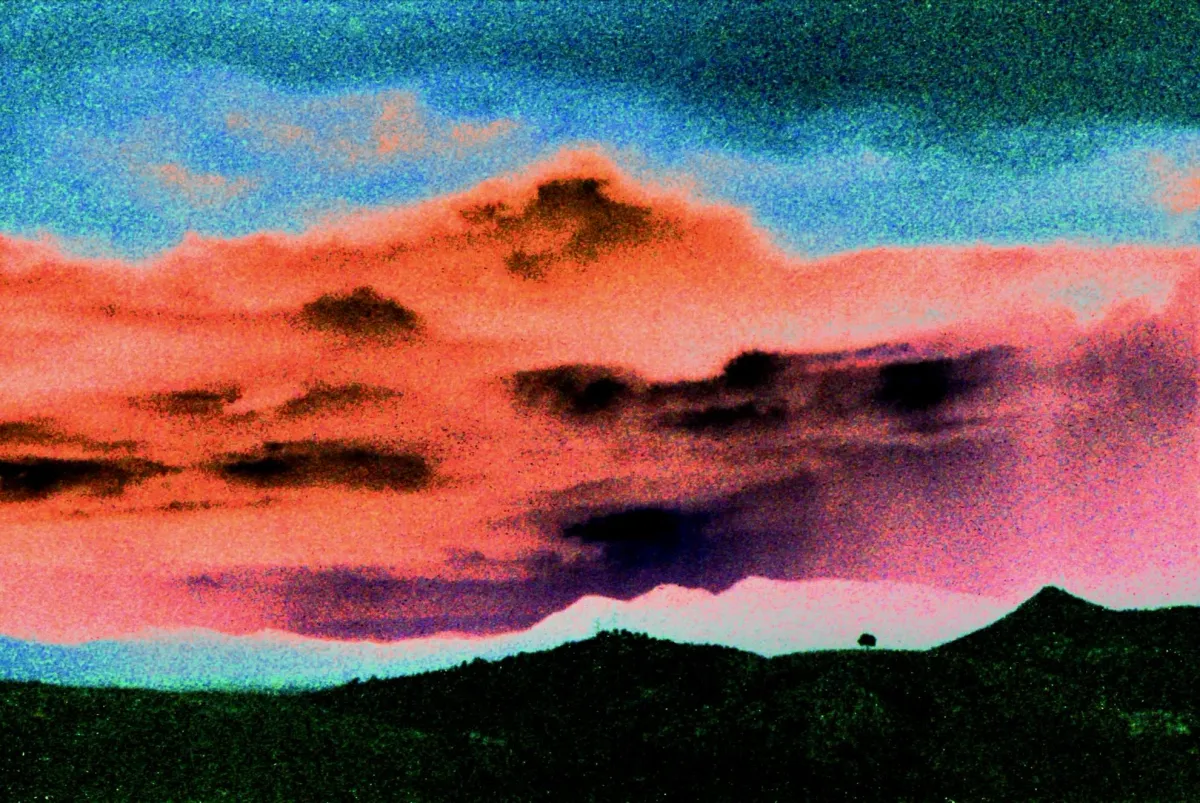 Photo of distant hills with a single, tiny, lost tree. The sky is dramatic orange, fuchsia, and turquoise. The image is so grainy it looks more like a painting than a phjoto.